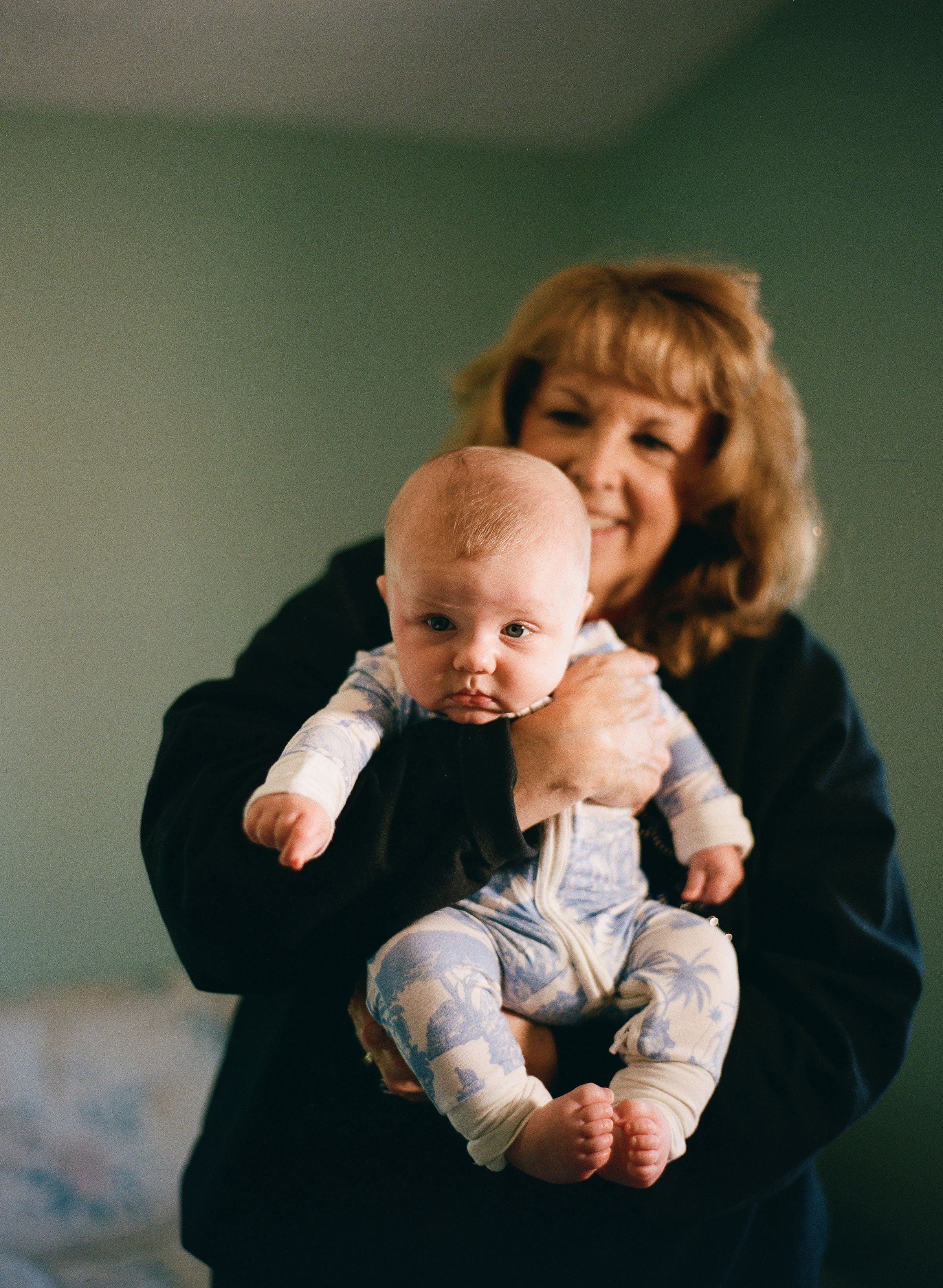 A smiling woman with blonde hair holds a baby in a blue floral onesie in a cozy indoor setting.