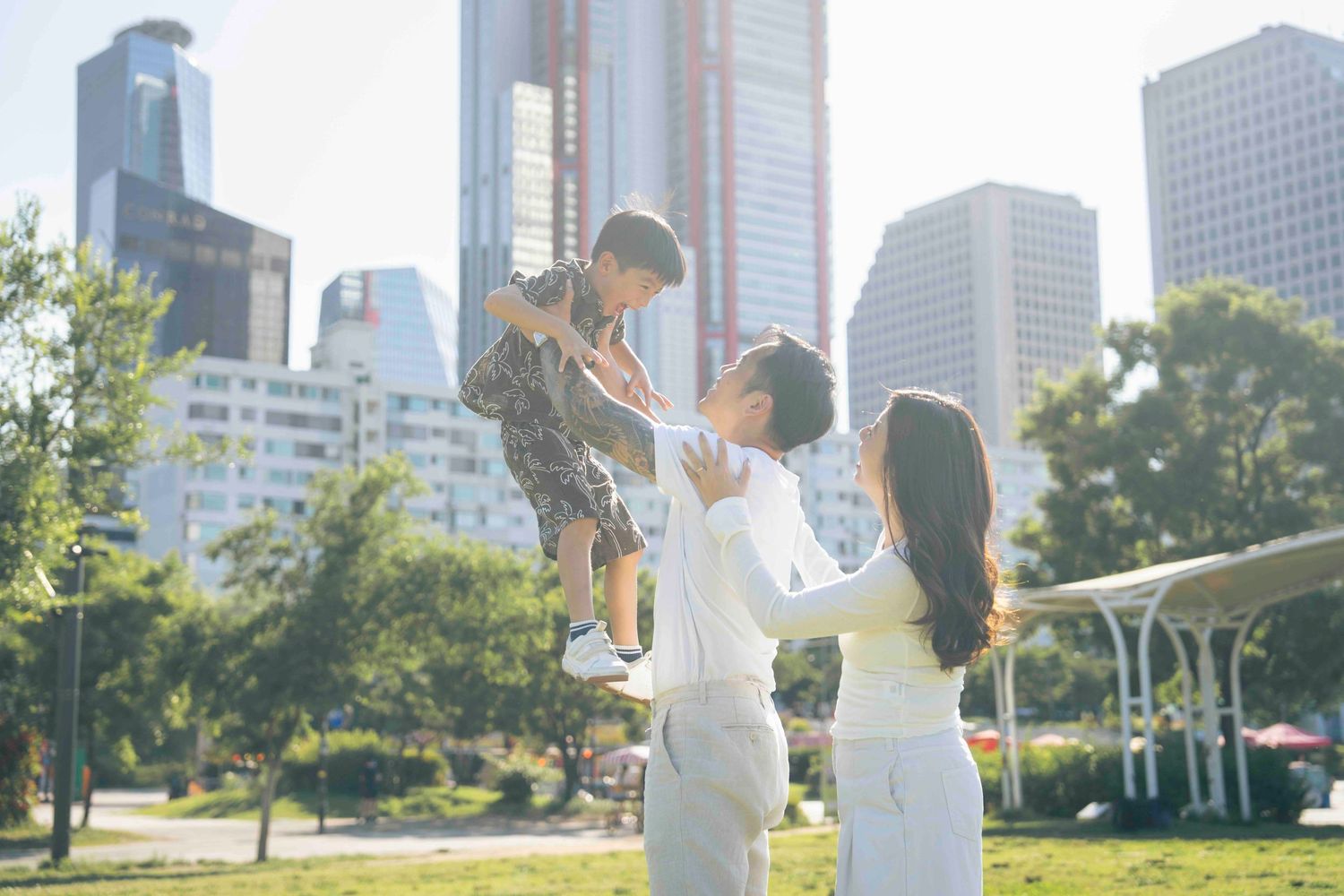 Family enjoying a sunny day in an urban park, parents lifting child with city skyscrapers in background.