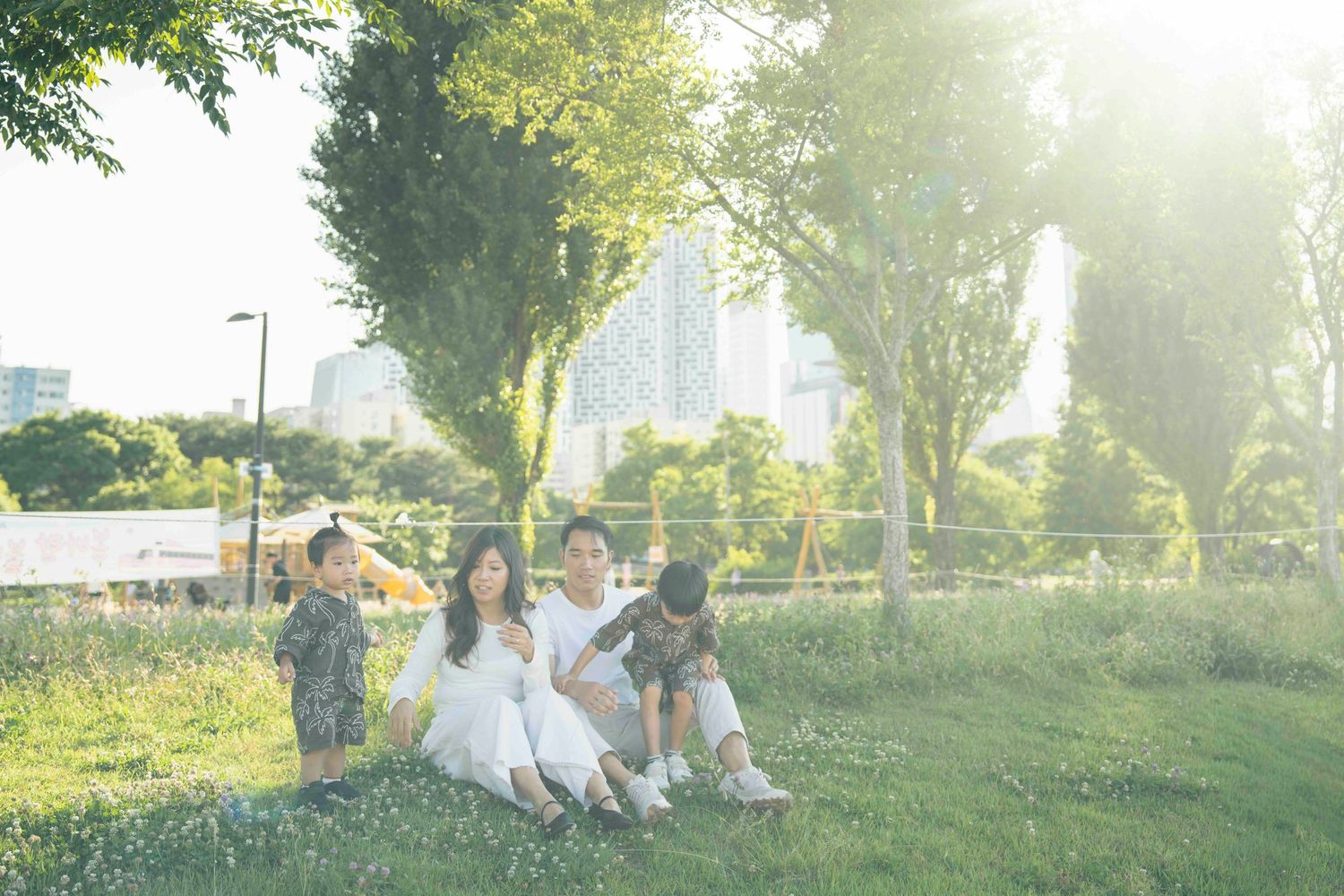 Family of four sitting on grass in a sunlit park with trees and city skyline in the background.