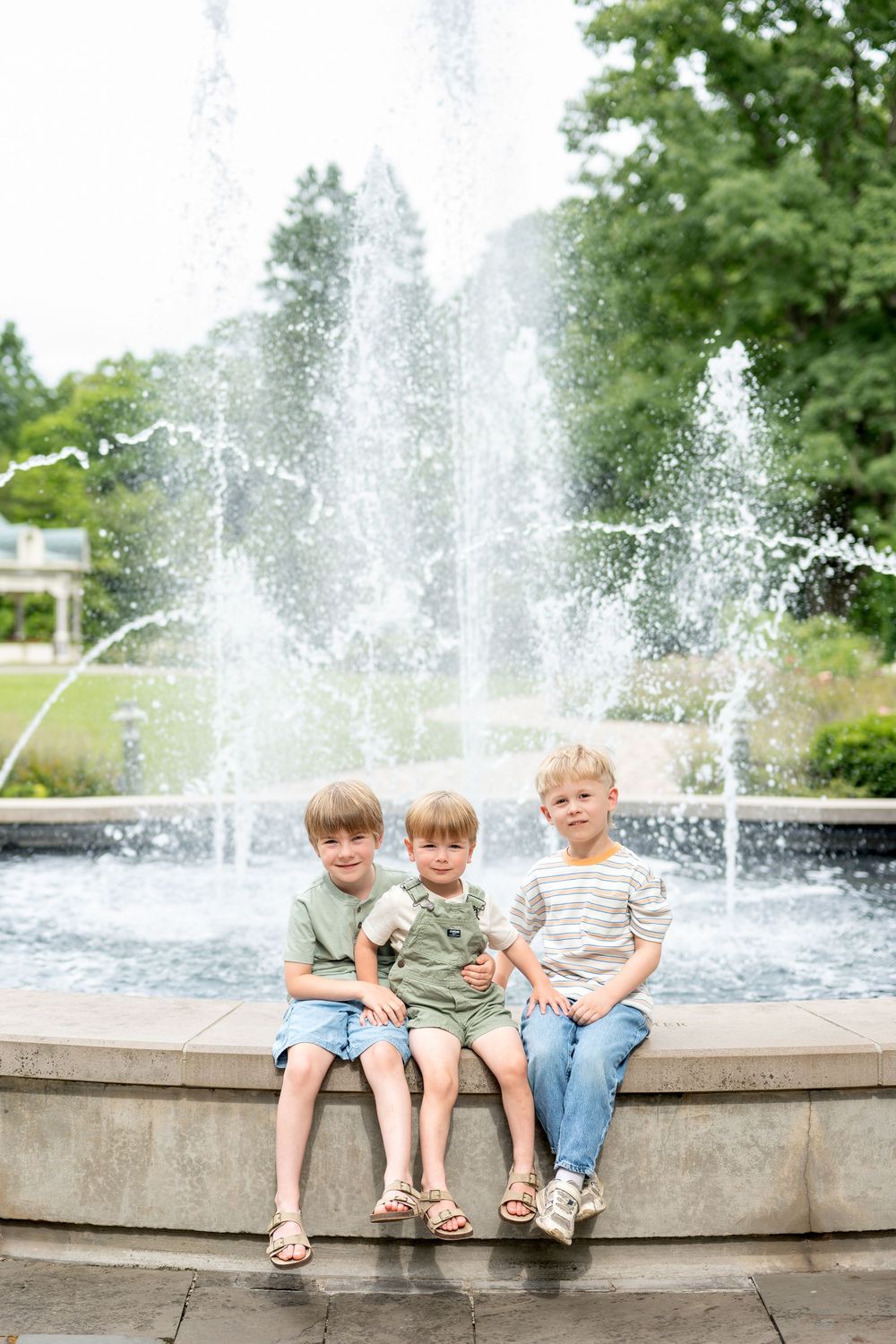 Three young boys sitting on fountain edge in a park, smiling together on a sunny day.