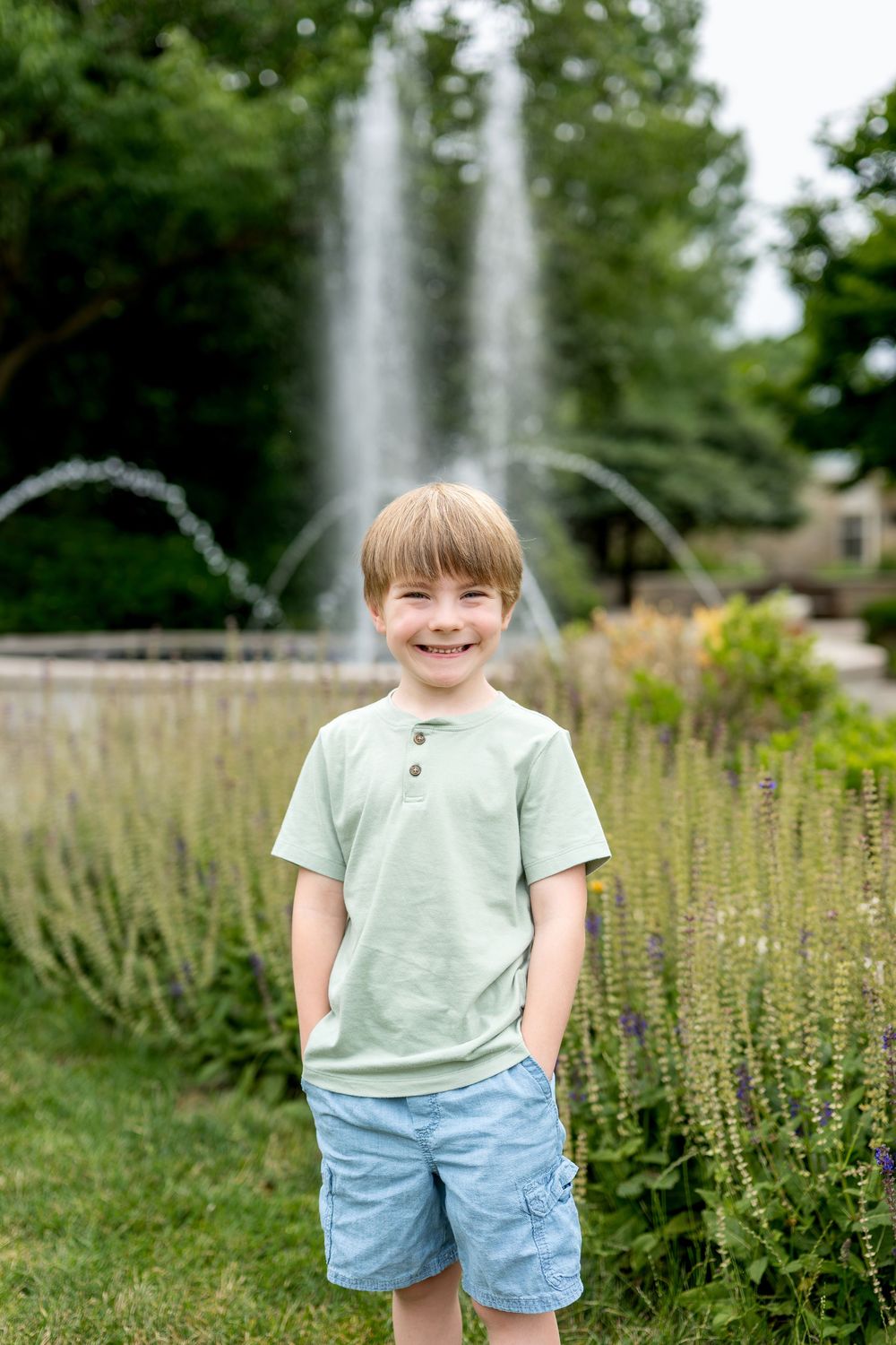 Smiling blonde boy in mint green shirt and blue shorts stands in front of a garden fountain on a summer day.
