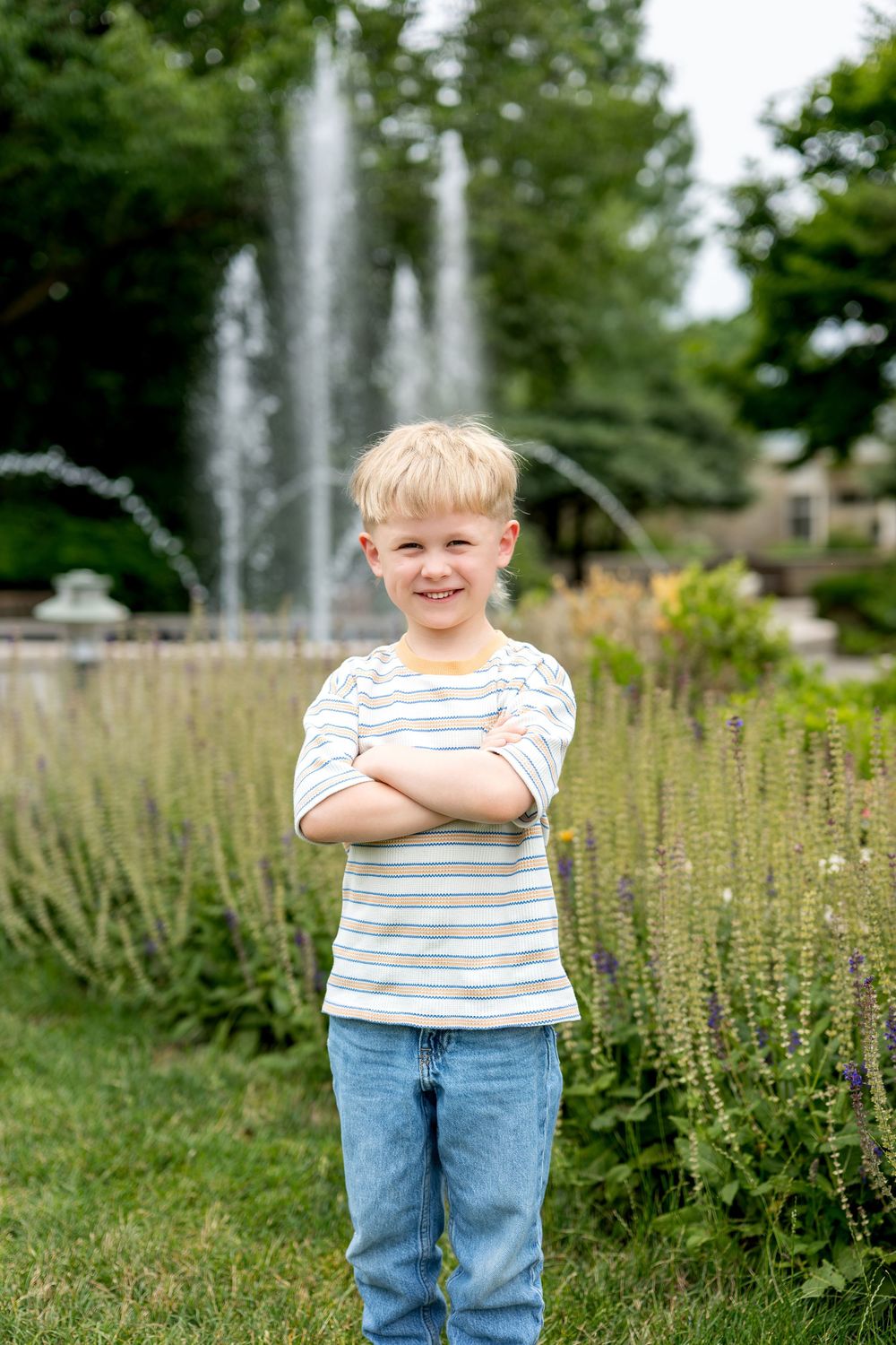 Smiling blond boy stands with arms crossed near a garden fountain.