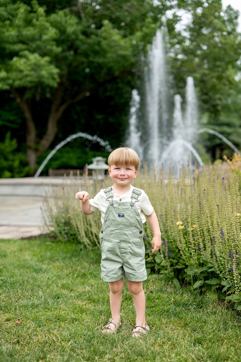 A smiling toddler boy in green overalls stands on grass near a garden fountain on a sunny day.