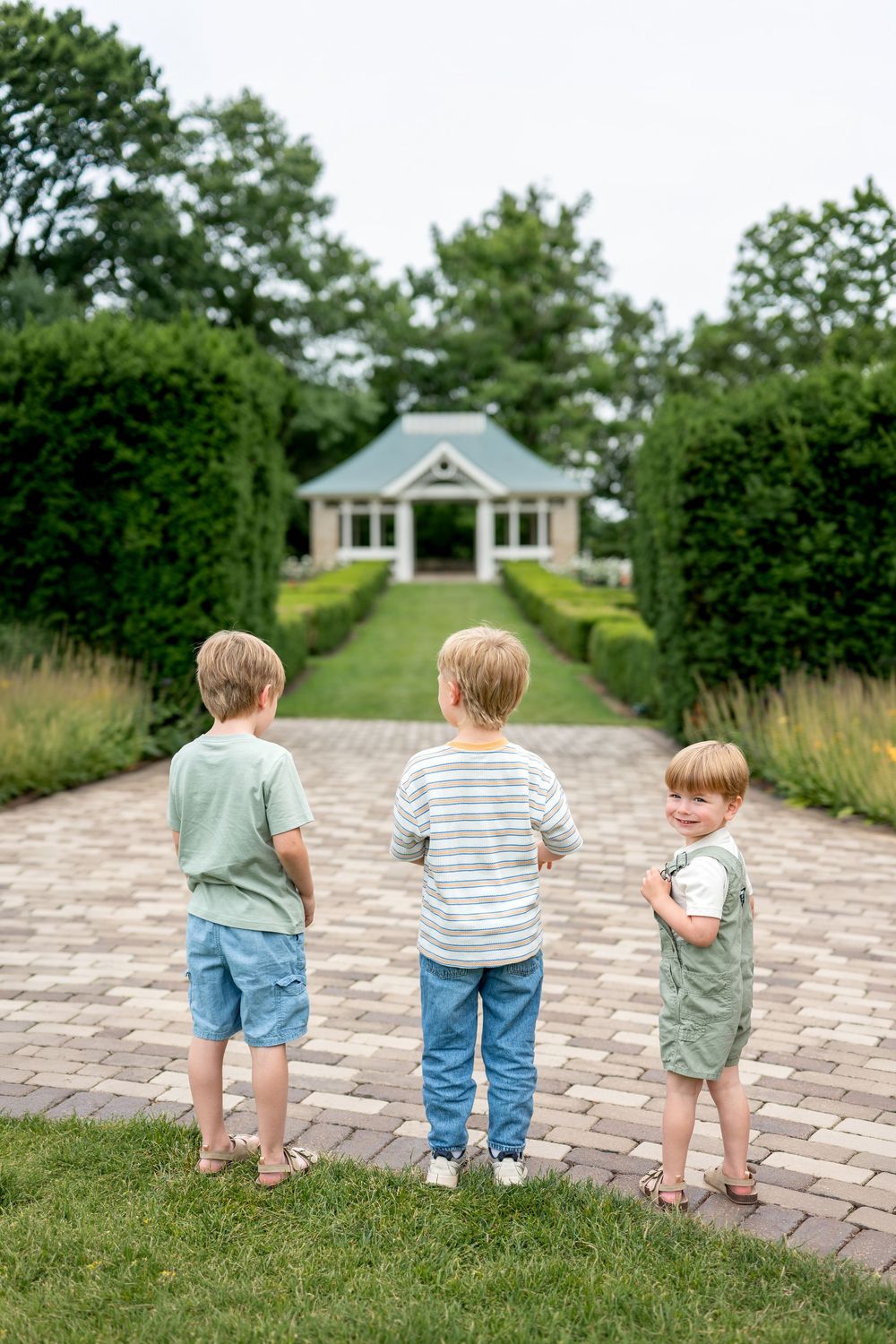 Three young boys stand on a brick garden path leading to a charming gazebo surrounded by lush green hedges.