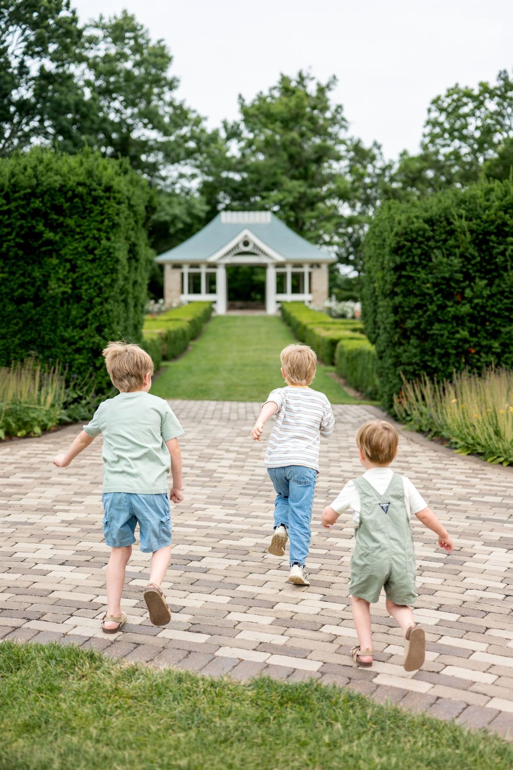 Three young boys run toward a charming garden gazebo on a brick path.