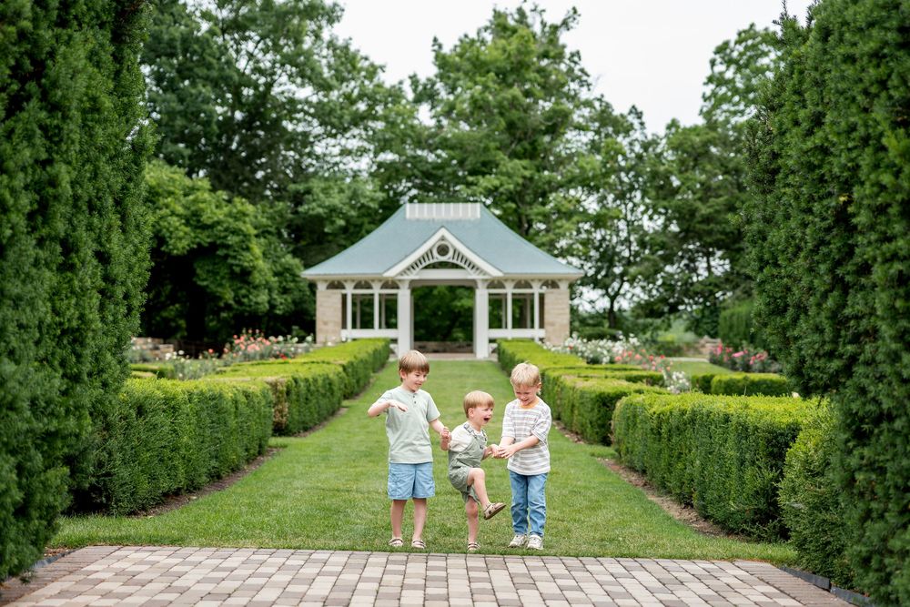 Three young children play together in a formal garden with a gazebo backdrop.