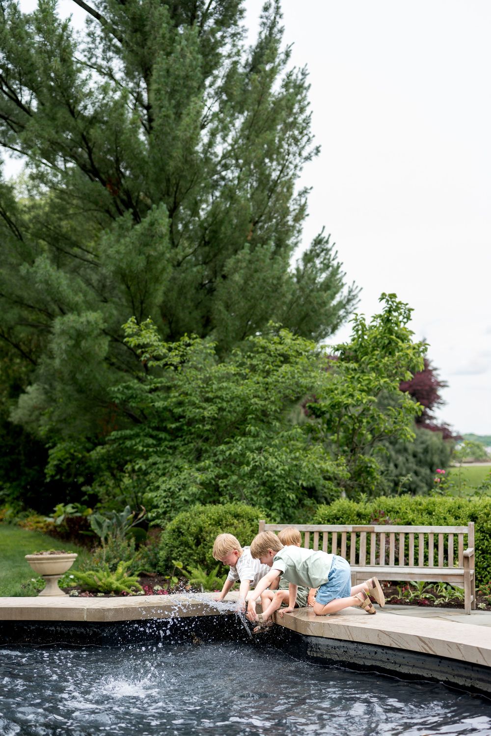 Two young children play at the edge of a decorative garden pond surrounded by lush greenery.