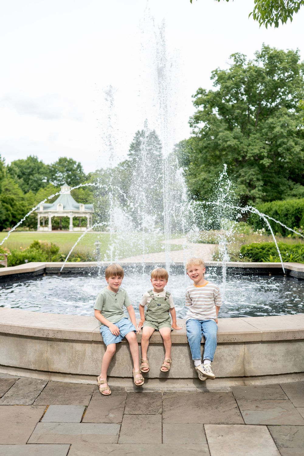 Three young children sit smiling on the edge of a decorative park fountain surrounded by lush green trees.