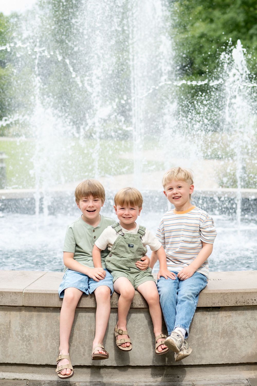 Three young boys sit smiling on a fountain ledge outdoors on a sunny day.