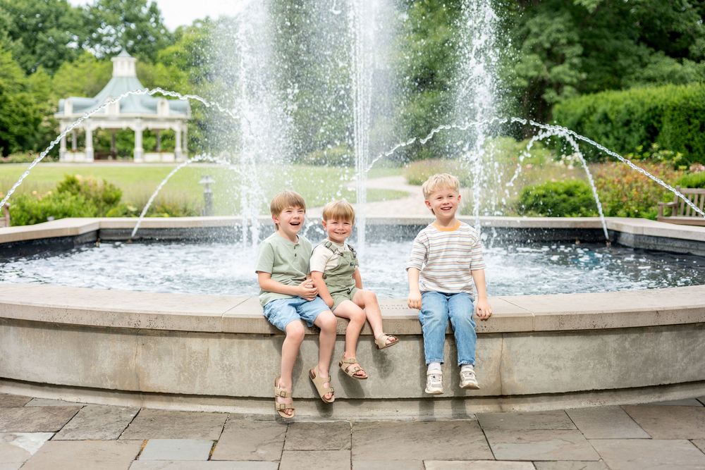Three young boys sit smiling on a stone fountain edge in a lush green park on a sunny day.