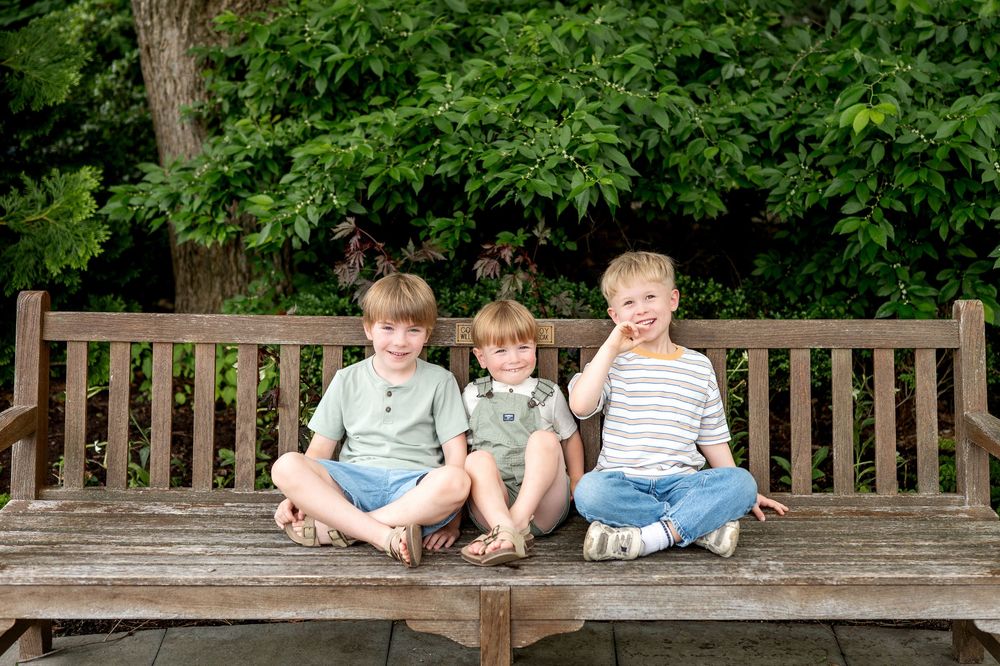 Three young boys sitting cross-legged on a wooden bench outdoors, smiling, surrounded by lush green trees.