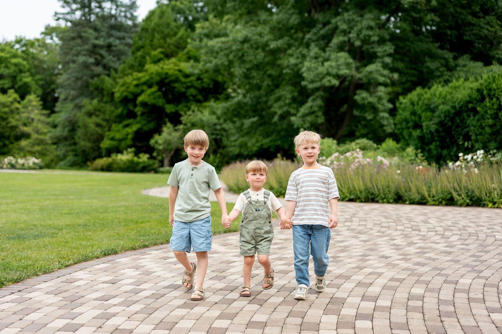 Three young boys holding hands and walking together on a brick path in a lush green park.