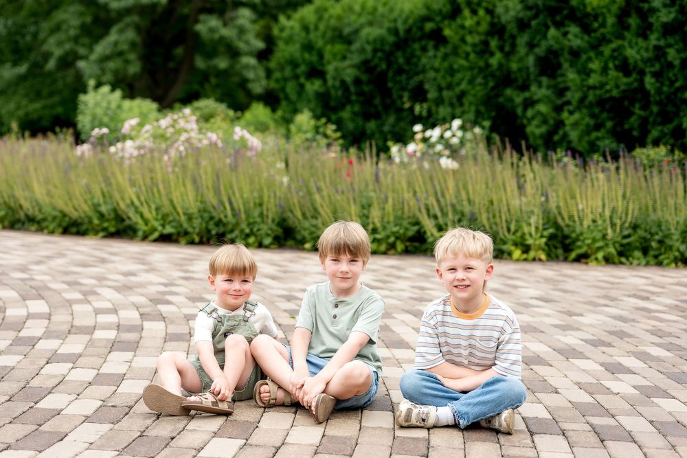 Three young boys sitting together on a brick patio surrounded by lush green gardens, smiling happily outdoors.