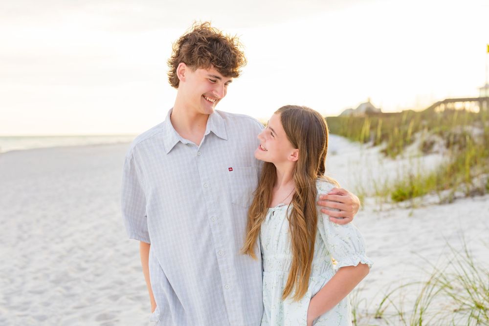A teenage boy and girl smile at each other on a sandy beach at sunset, the boy's arm around the girl.