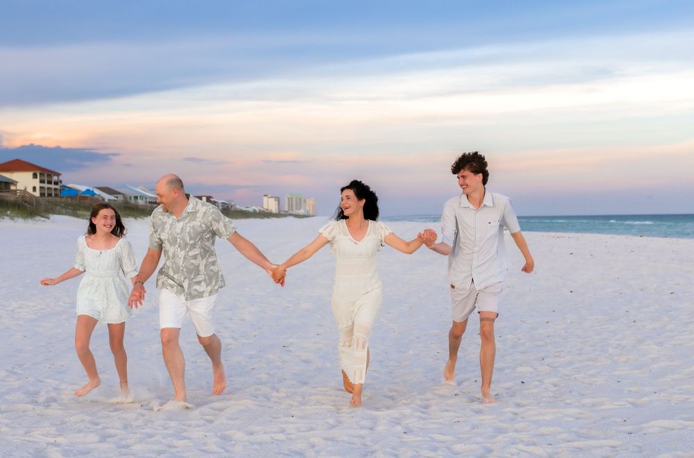 Family of four holding hands and jumping on a white sand beach at sunset wearing coordinated white outfits.