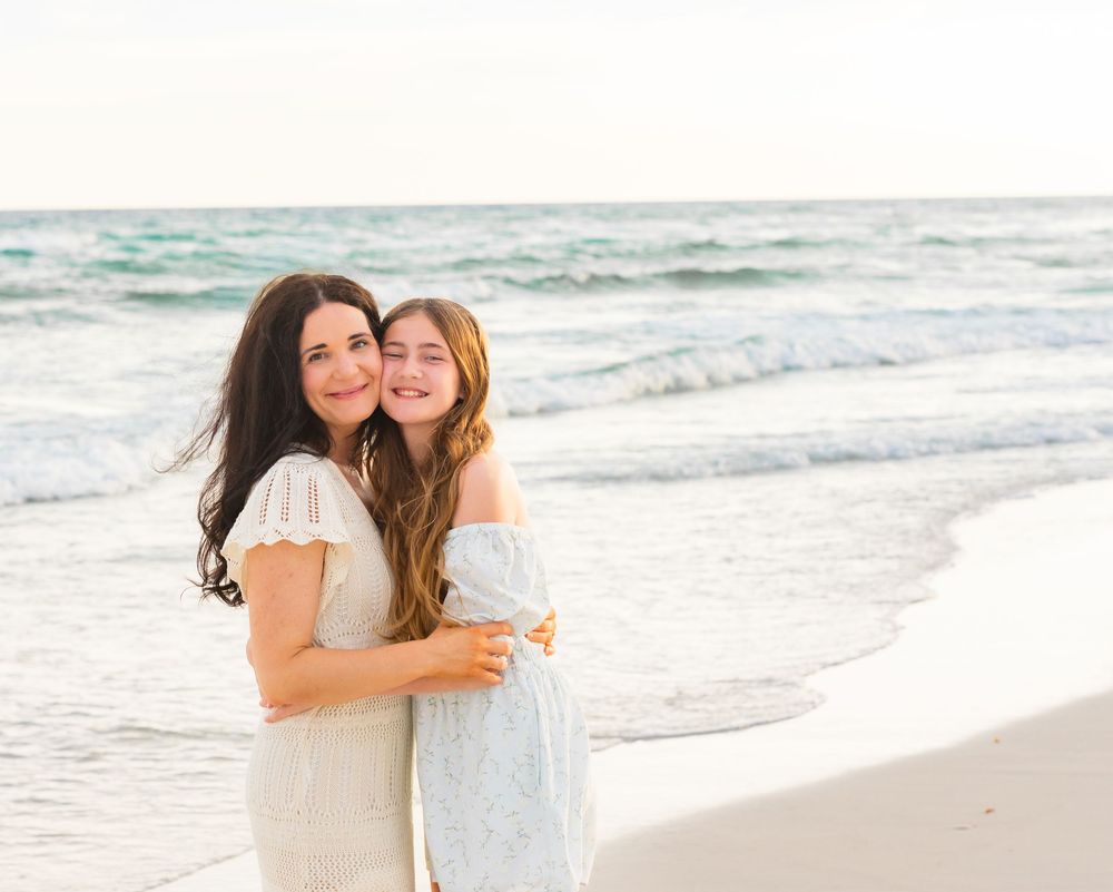 Two women in white dresses embracing and smiling on a sunny beach with ocean waves behind them.