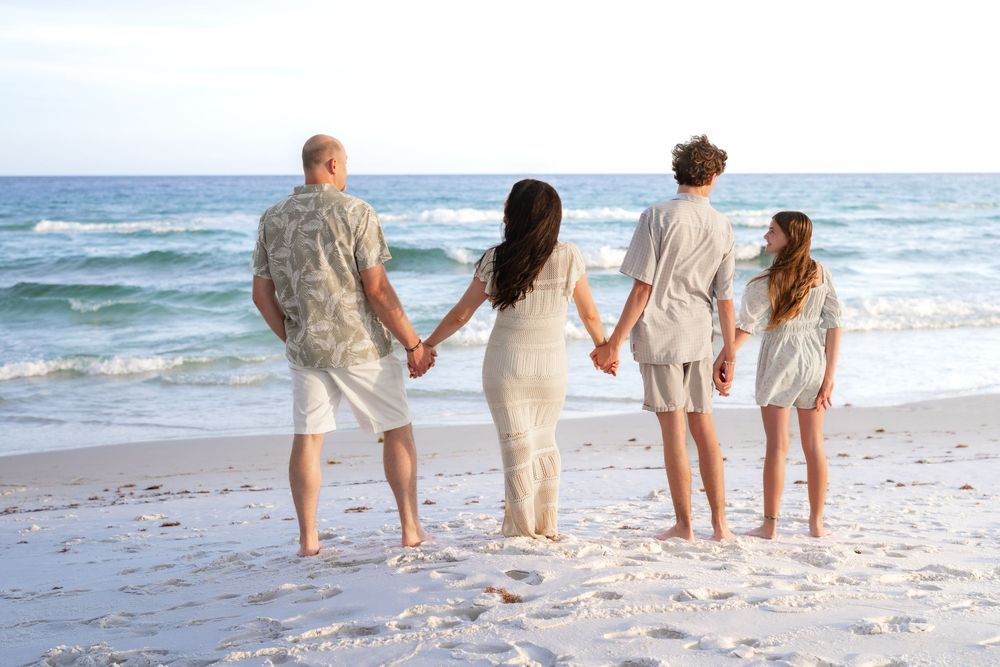 Family of four holding hands on a sandy beach, facing ocean waves on a sunny day.