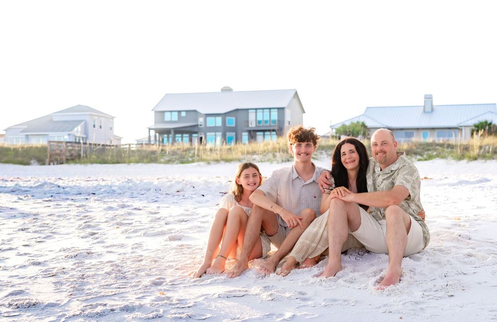 Family of four sitting together on white sand beach with beach houses in the background on a sunny day.