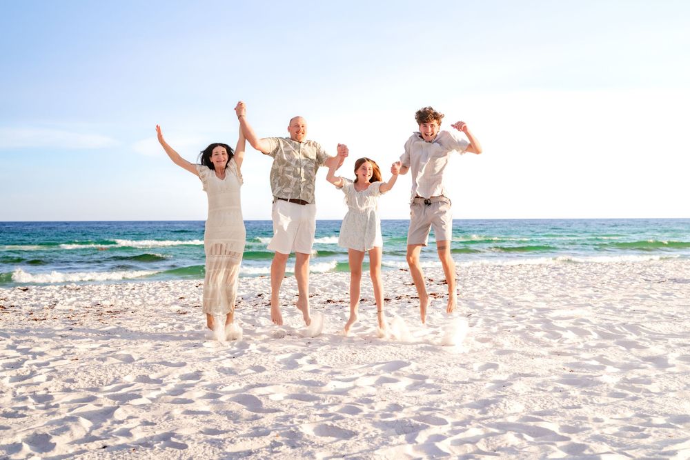 Joyful family of four jumping in the air on a sunny white sand beach with blue ocean waves behind them.