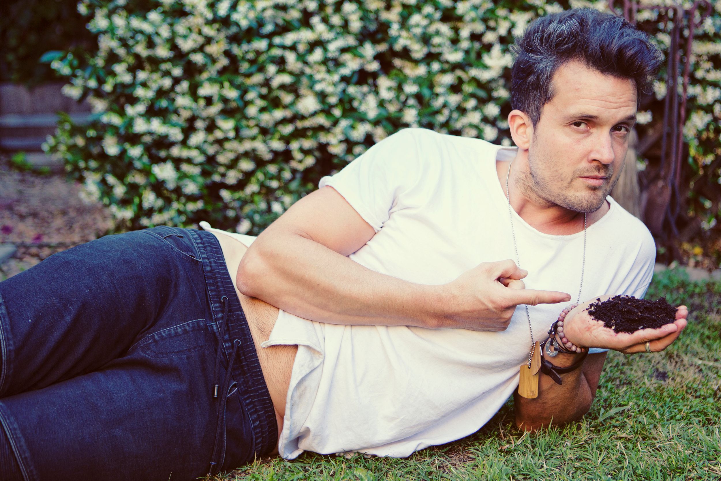Man in white t-shirt and jeans lying on grass, pointing at soil in his hand, with green hedges behind.