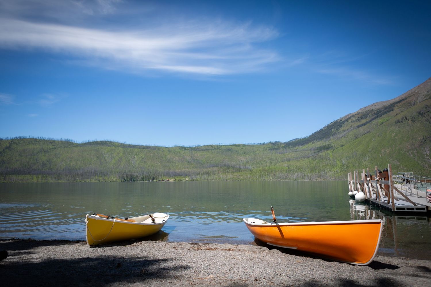 Two colorful rowboats on a rocky lakeshore with calm water, green hills, and a wooden dock under a blue sky.