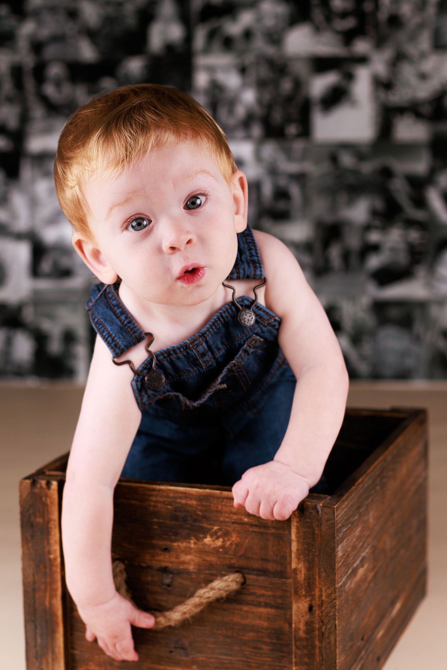 Adorable red-haired baby in denim overalls sitting inside a rustic wooden crate against a photo collage backdrop.