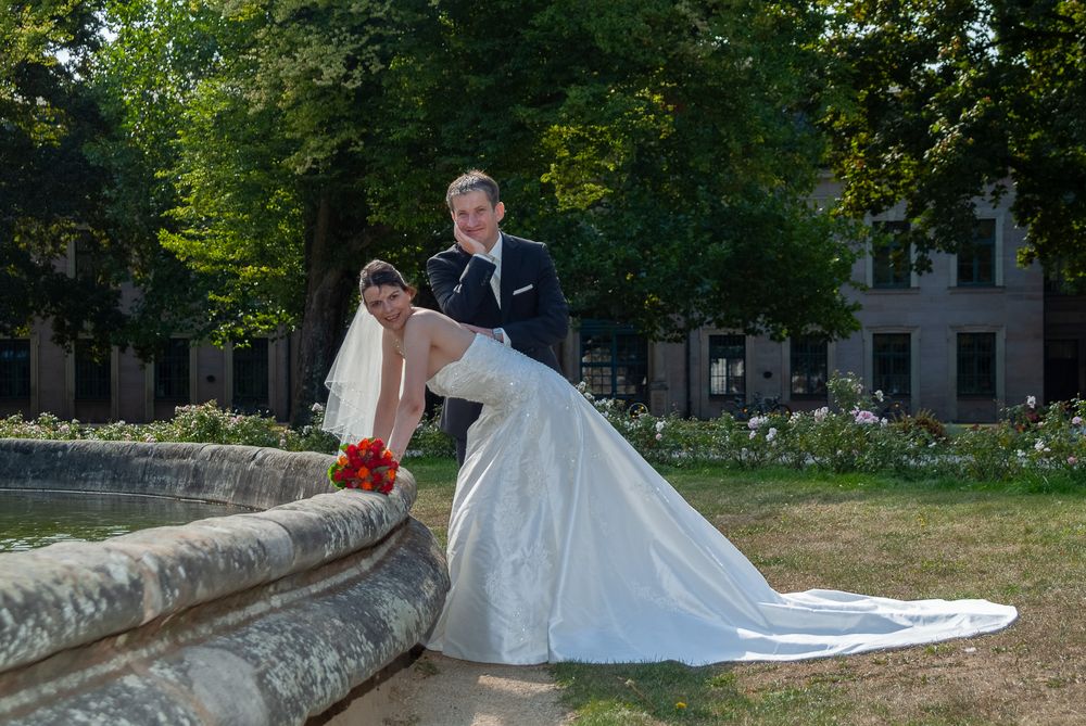 Braut sitzt elegant am Rand eines Steinbrunnens, der Bräutigam steht bei ihr. Klassische Hochzeitsbilder im Stadtpark Erlangen.