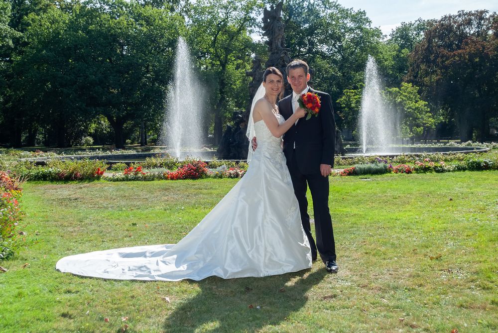Brautpaar posiert elegant vor zwei Springbrunnen im Stadtpark Erlangen. Professionelle Hochzeitsfotografie mit traumhaftem Brautkleid.