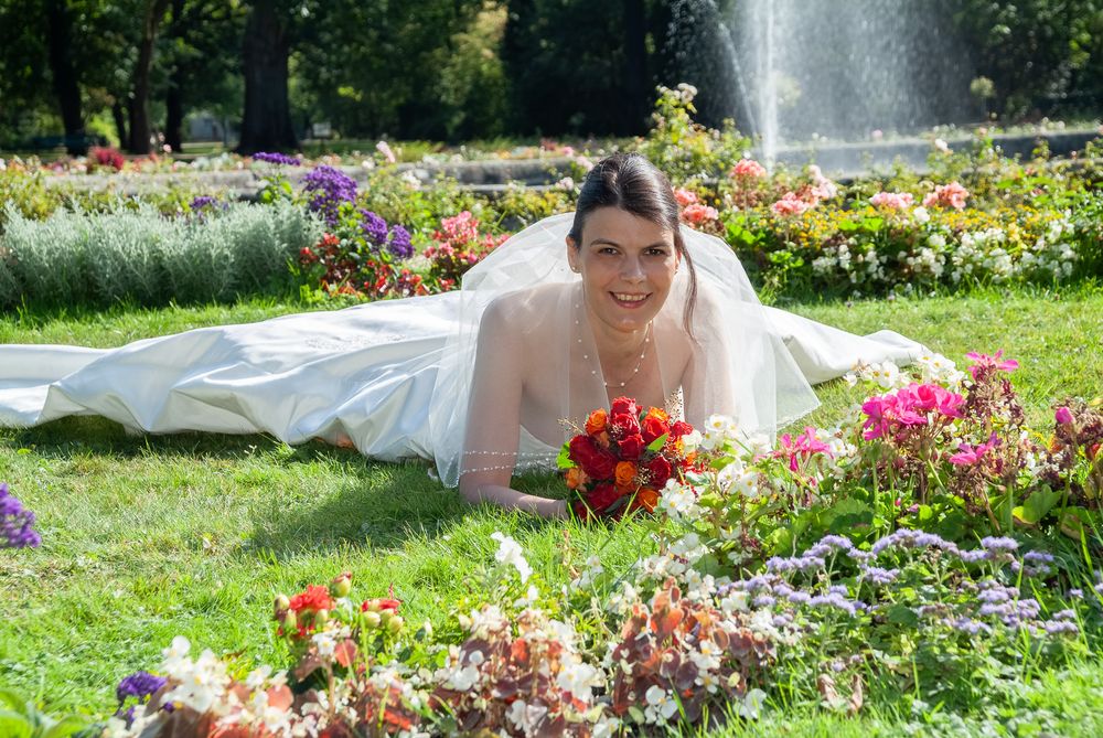 Braut liegt elegant auf der Wiese vor bunten Blumenbeeten und Springbrunnen. Kreatives Brautporträt im Stadtpark Erlangen.