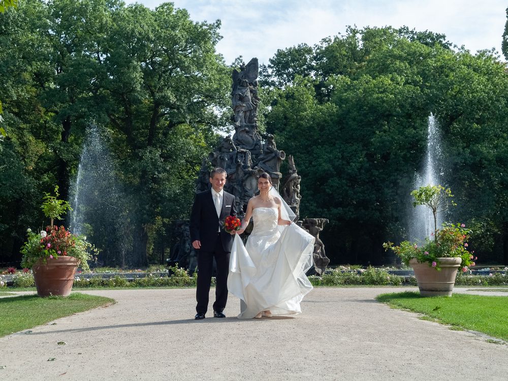 Brautpaar spaziert Hand in Hand durch den Stadtpark Erlangen mit Springbrunnen im Hintergrund. Weitläufige Hochzeitsfotografie.