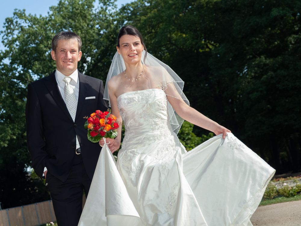 Strahlendes Brautpaar posiert im Grünen, die Braut rafft ihr weißes Kleid. Professionelle Hochzeitsfotografin in Erlangen.