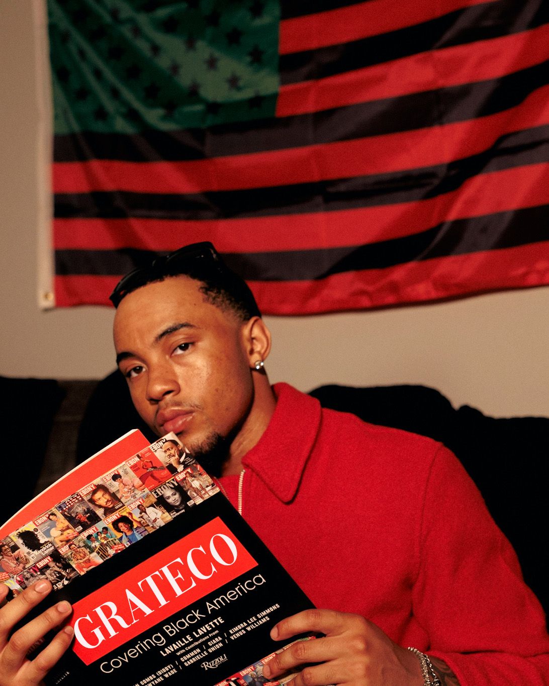 Man in red jacket holding GRATECO Covering Black America magazine in front of a red, black, and green flag.