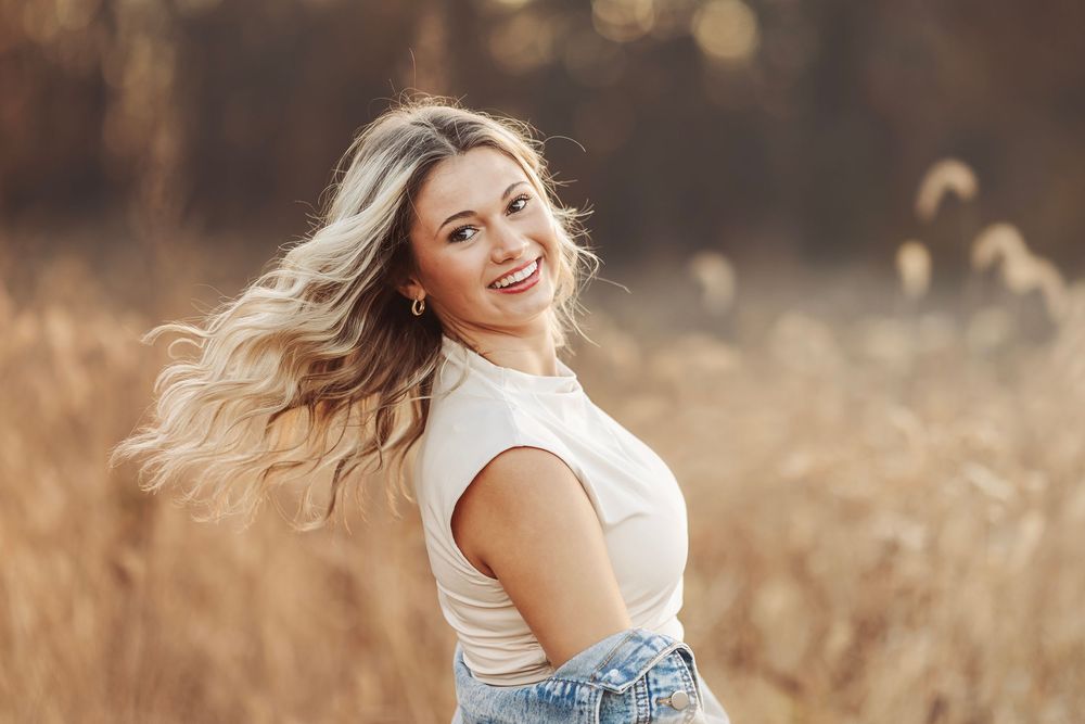 Young woman in black crop top and jeans smiling, posing against a wall outdoors in a sunny setting.