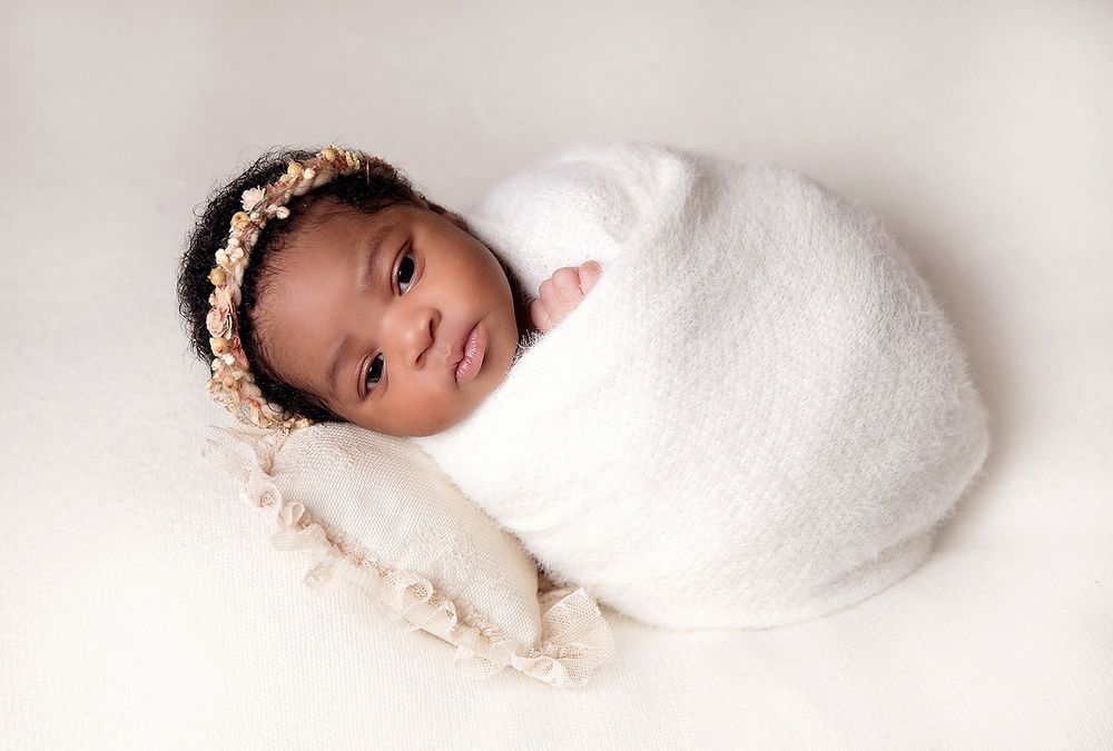 Newborn baby girl swaddled in white wrap with floral headband, resting on soft pillow in studio photo. during a newborn session in Bexley