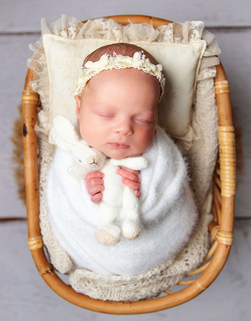 Newborn baby swaddled in white wrap holding a knit bunny toy, sleeping in a bassinet with lace trim. During a newborn photos in Bexley.