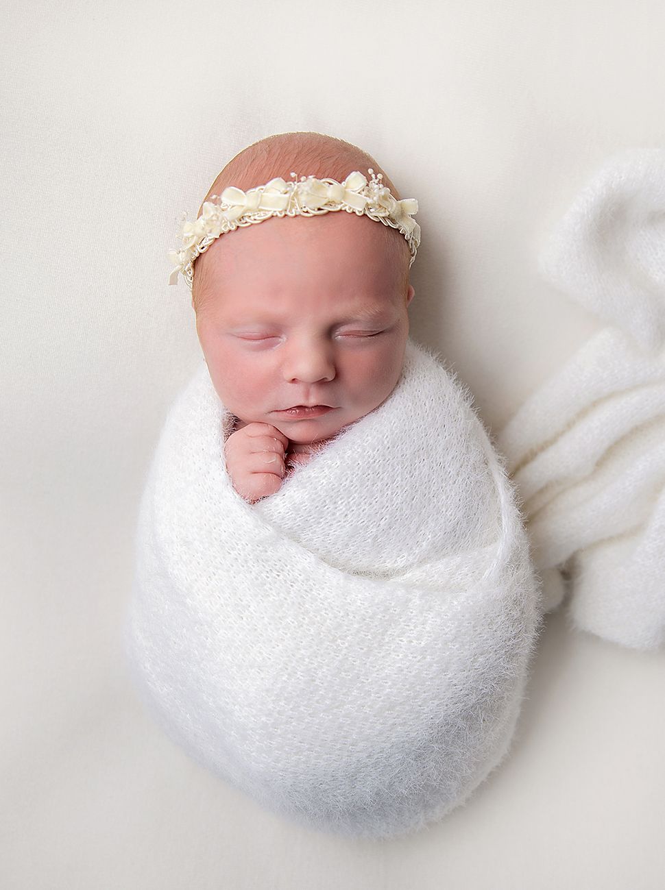 Sleeping newborn baby swaddled in white wrap wearing a floral headband, photographed on white background at a Bexley newborn photoshoot