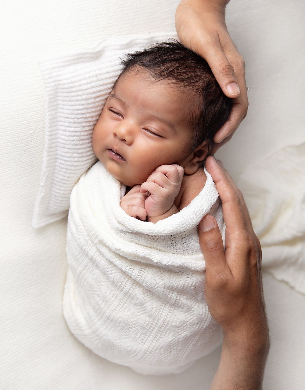 Baby swaddled in white wrap sleeping peacefully while adult hands gently cradle the infant's head. During a newborn session in Bexley.