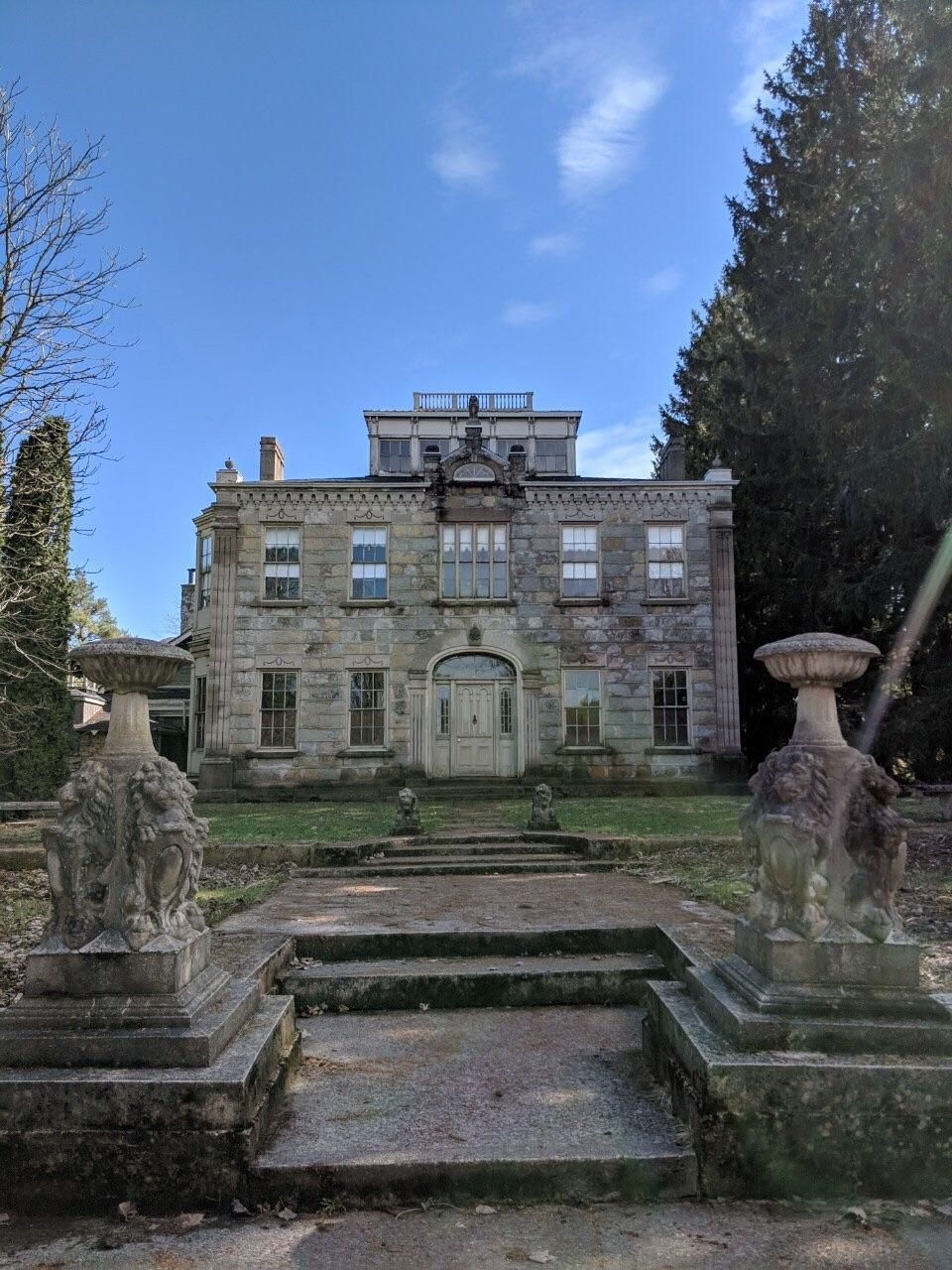 Historic stone manor house with ornate statues and stone urns flanking steps leading to grand entrance under blue sky.