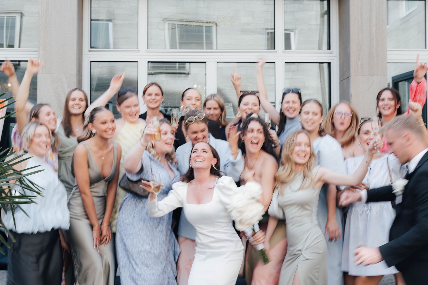Bride in white gown celebrates with large bridal party in elegant dresses outside modern building.