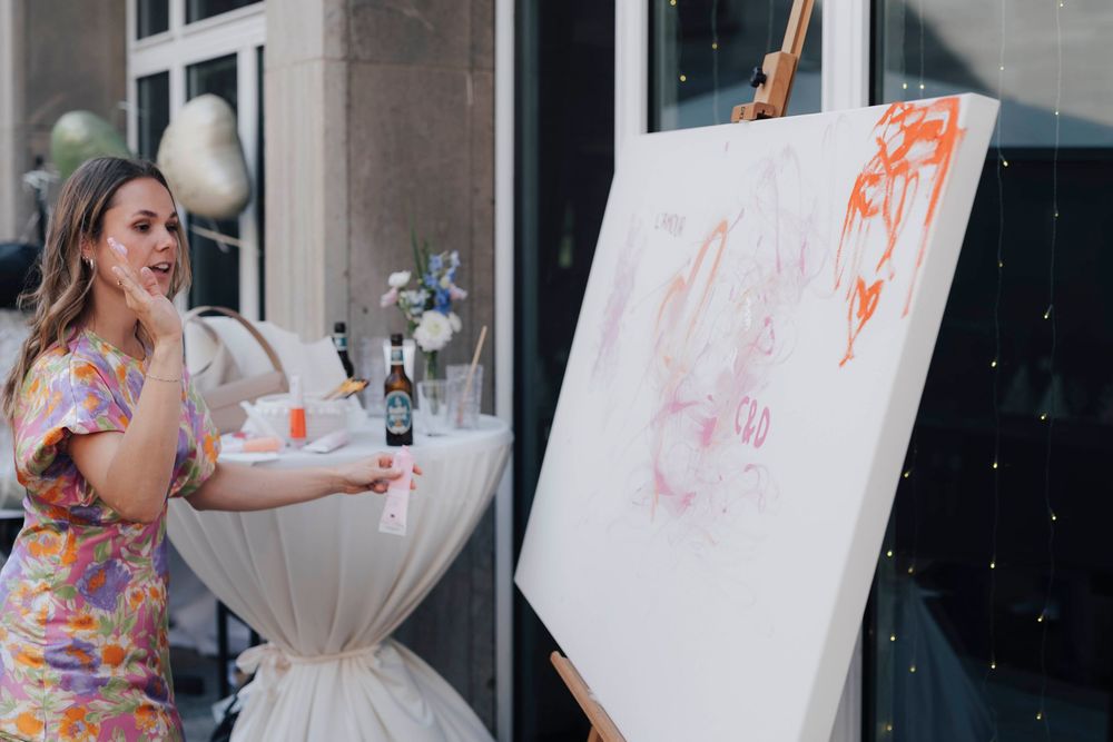 Woman in floral dress painting on large canvas at easel during outdoor art event.