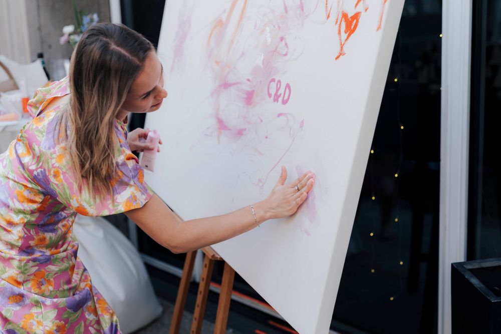 Woman in floral dress painting on large white canvas on easel at indoor art event.