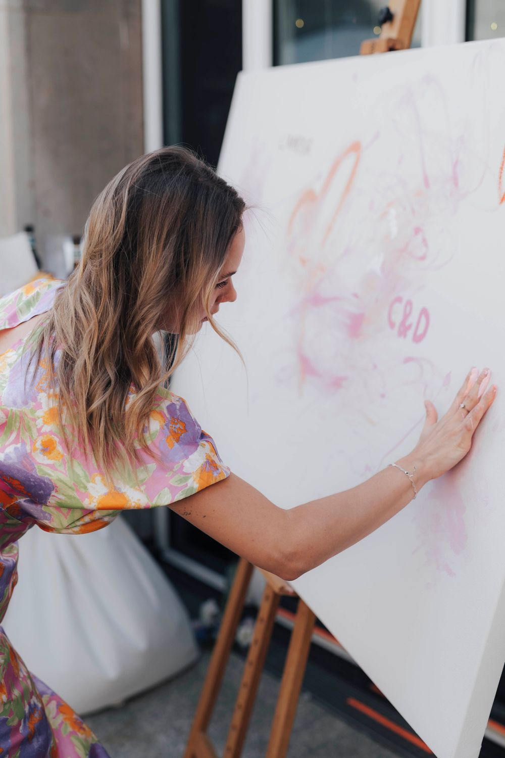 Woman in floral dress painting on large canvas on easel, creating pink abstract artwork in outdoor art studio.