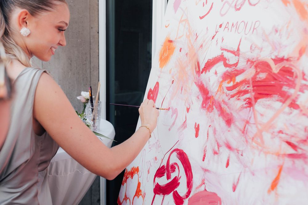 A smiling blonde woman in a silver dress creates a vibrant pink and red abstract painting on a large canvas.