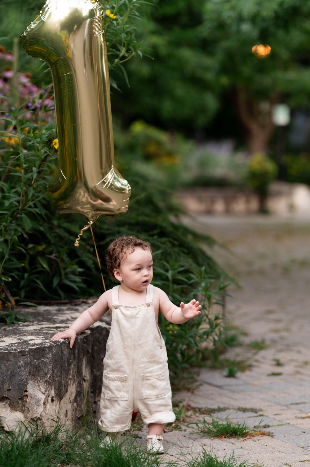 Toddler in cream overalls stands by stone wall holding gold number 1 balloon in lush garden.