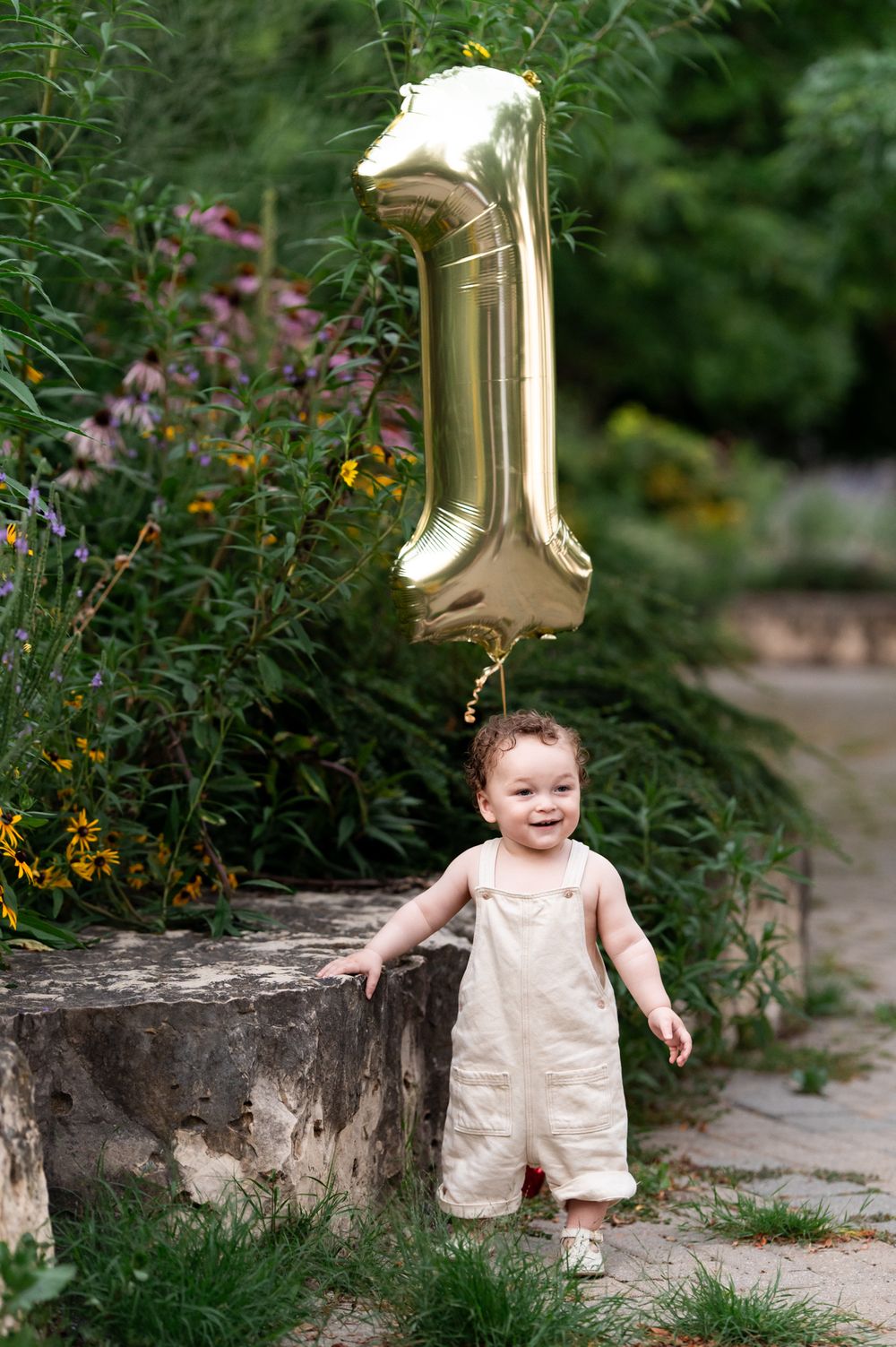 A smiling baby in cream overalls stands outdoors holding a gold number 1 balloon, celebrating their first birthday.