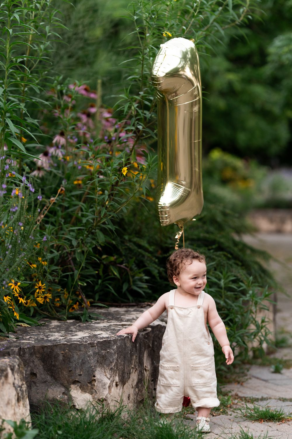Happy toddler in cream overalls smiles holding gold number 1 balloon beside stone wall in garden.