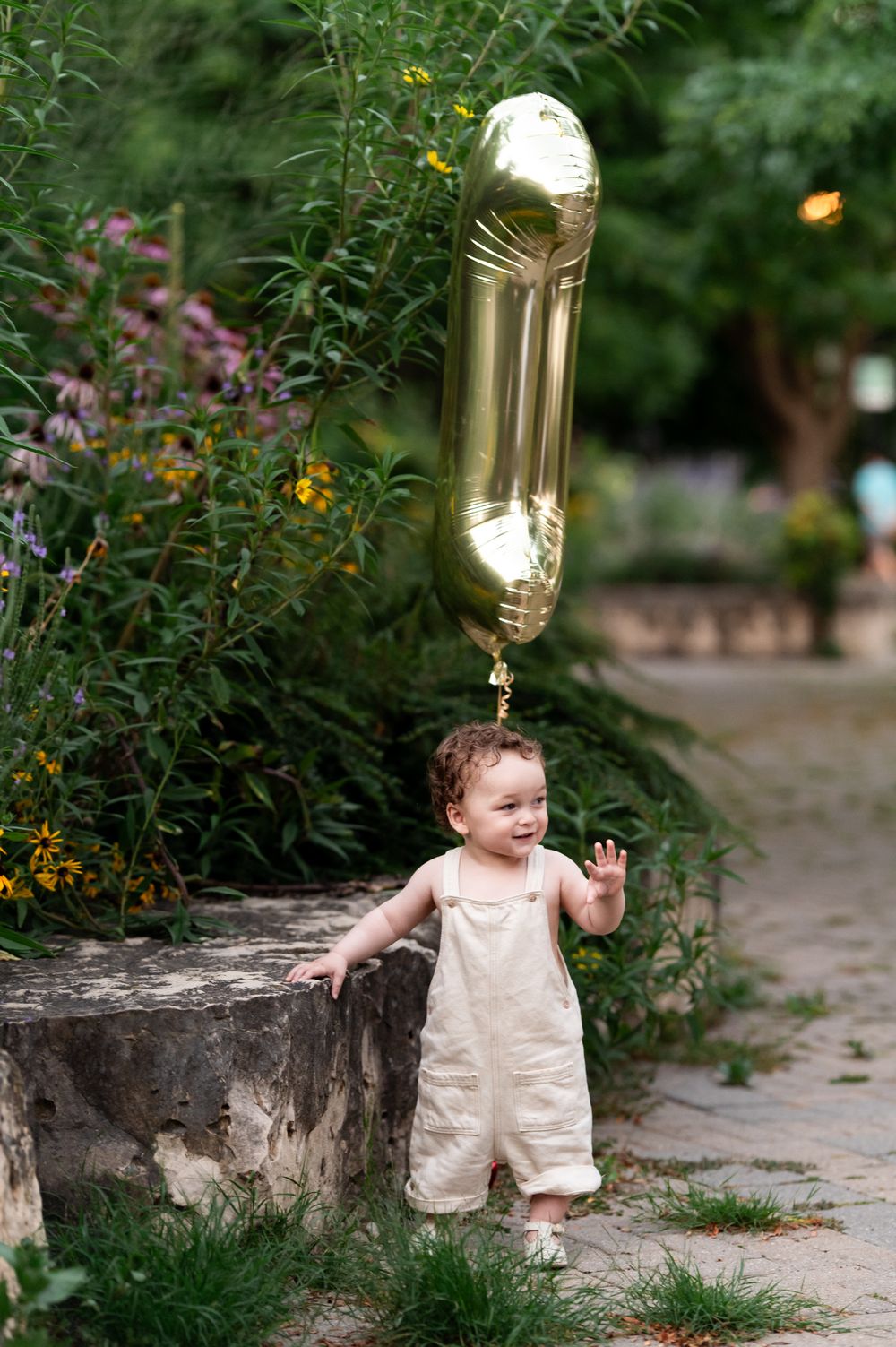 Toddler in cream overalls holding a gold number one balloon standing by a stone planter in a lush green park.