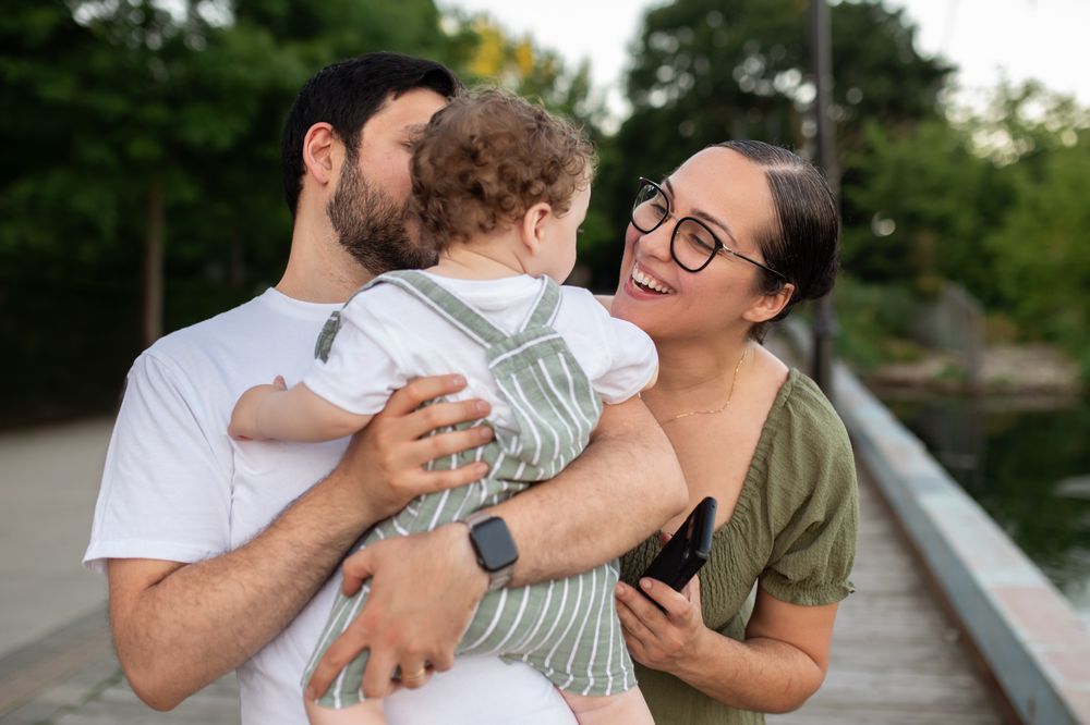 A happy young family with a baby outdoors, parents smiling and holding their toddler on a wooden bridge.