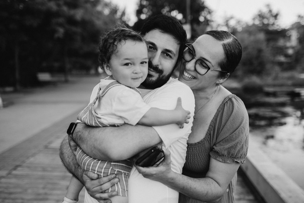 Black and white photo of a happy young family, parents smiling and holding their toddler outdoors near a park.
