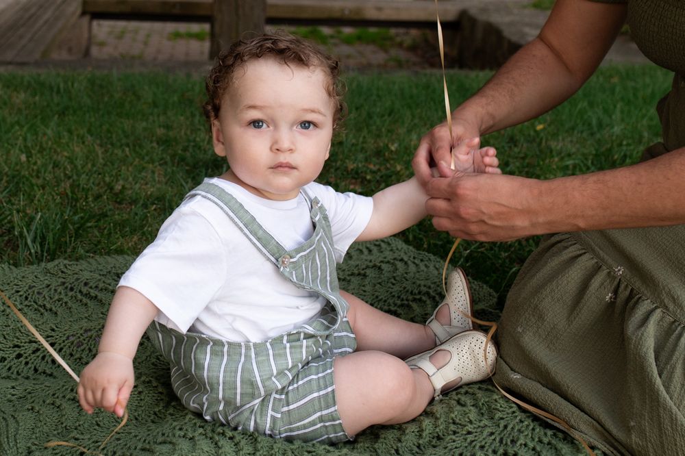 Adorable baby in striped overalls sitting on green blanket outdoors, holding a straw with an adult nearby.